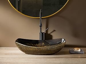 Lavabo de baño ovalado negro y dorado con grifo alto. Espejo redondo con borde dorado sobre encimera de madera rústica.