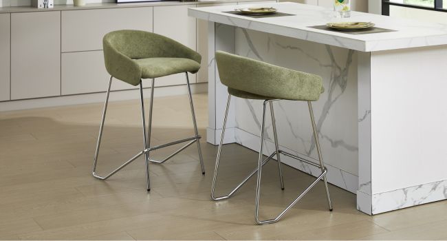 Salle à manger moderne avec deux chaises rembourrées vert sauge avec des bases en fil de fer chromé positionnées sur un comptoir ou une table blancs contre un mur blanc.