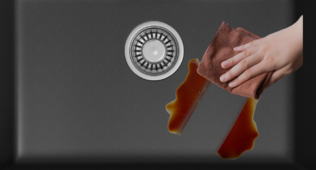 Sink drain with hand using reddish-brown sponge or cloth to clean dark surface, possibly demonstrating cleaning product or technique.