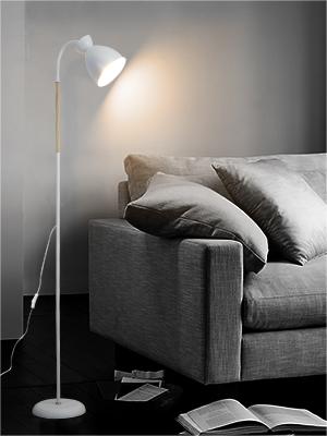 Weiße Stehlampe, die eine graue Couch mit Kissen beleuchtet. Ein offenes Buch auf dem Boden neben der Lampe in einem schwach beleuchteten Raum