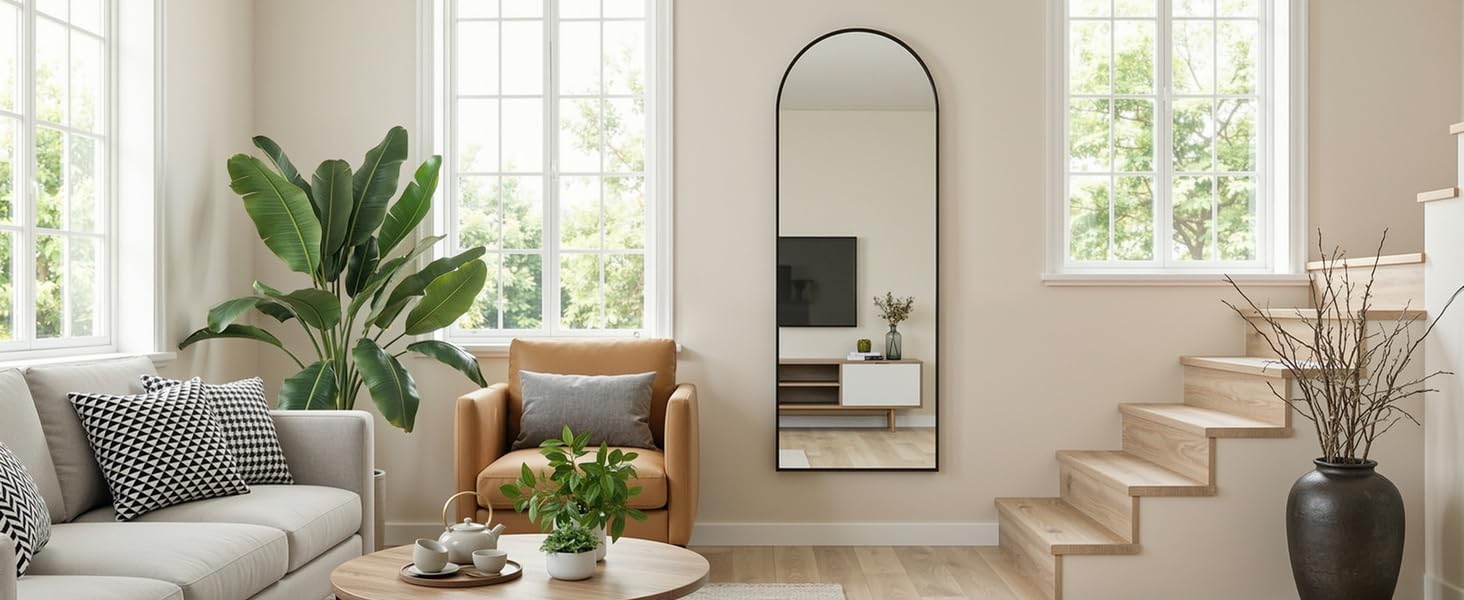 Modern living room interior with large arched mirror, gray sofa, potted plants, and bright natural lighting through windows.