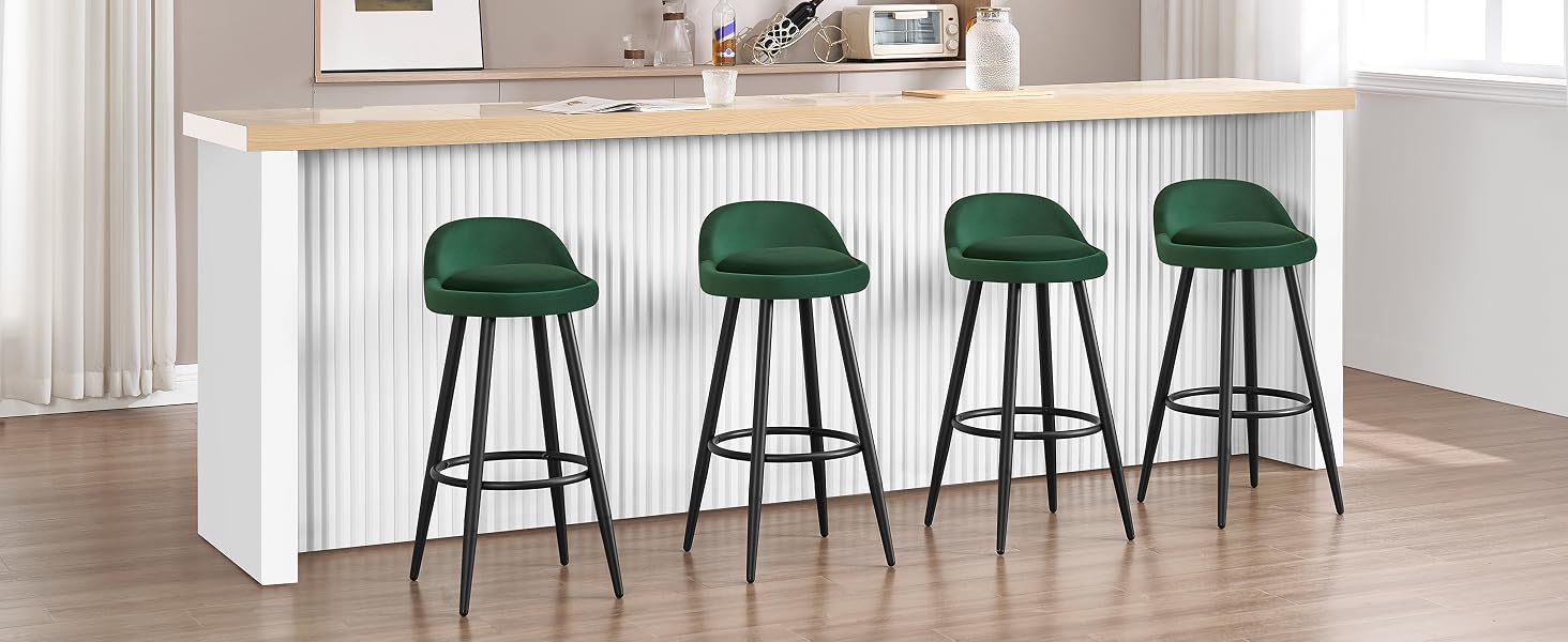 Cuatro taburetes de bar de terciopelo verde con patas de metal negro dispuestos en una isla de cocina blanca con una encimera de madera. Pequeños electrodomésticos visibles en el mostrador