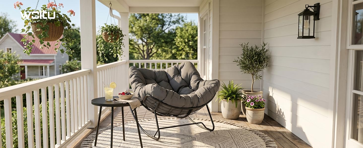 Chaise papasan noire avec coussin rond sur porche blanc avec balustrades, plantes en pot et verdure suspendue visibles en arrière-plan.