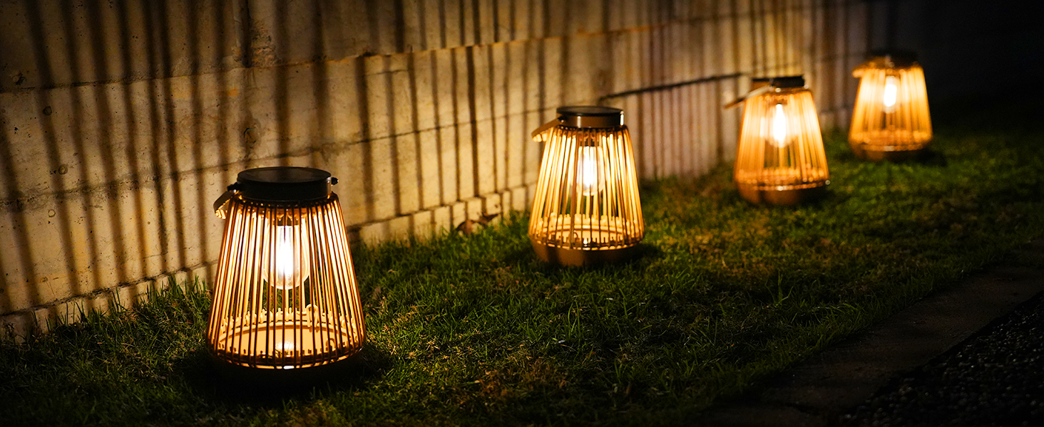 Quatre lanternes solaires extérieures avec des cages en bambou qui brillent chaleureusement sur l'herbe à côté d'une clôture. Les lanternes créent une ambiance chaleureuse en soirée