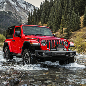 Jeep Wrangler rossa che attraversa un fiume o un torrente, dimostrando capacità fuoristrada in un ambiente forestale montuoso. Sono mostrati diversi angoli di guado.