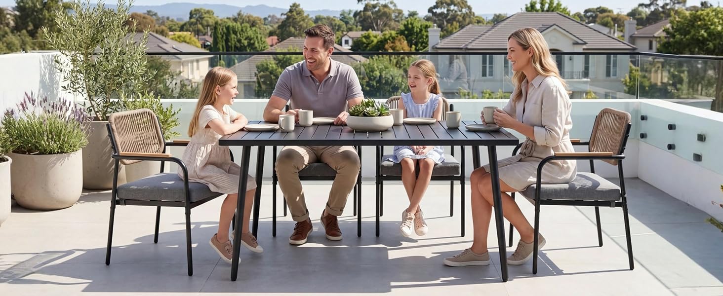 Scène de repas en plein air avec la famille assise à une table en bois et des chaises en métal noir sur une terrasse pavée, entourée de plantes en pot et d'un jardin naturel à la lumière du jour.