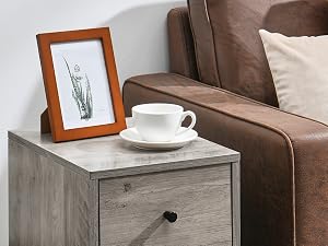 Grauer Nachttisch aus Holz mit drei Schubladen neben einem braunen Ledersessel. Dekoriert mit einem gerahmten Bild, einer Kaffeetasse und einer Pflanze im Hintergrund
