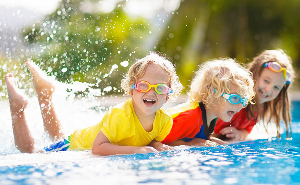 Des enfants portant des lunettes de natation colorées jouent en eau peu profonde. Des éclaboussures d'eau les entourent lorsqu'ils sont couchés sur une surface bleue, probablement dans une piscine ou une aire de jeux aquatiques