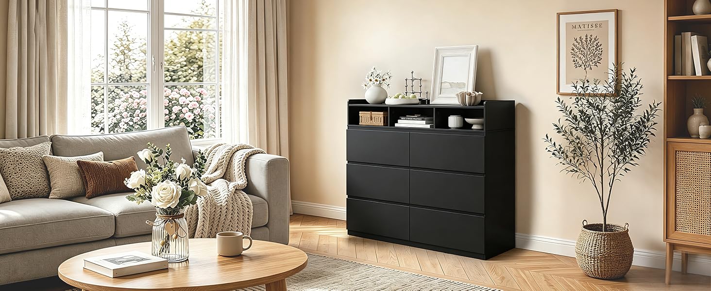 Comò nero a tre cassetti con hardware dorato esposto in un soggiorno moderno con divano beige, specchio rotondo, pianta in vaso, finestra, libreria e decorazioni dai toni neutri.