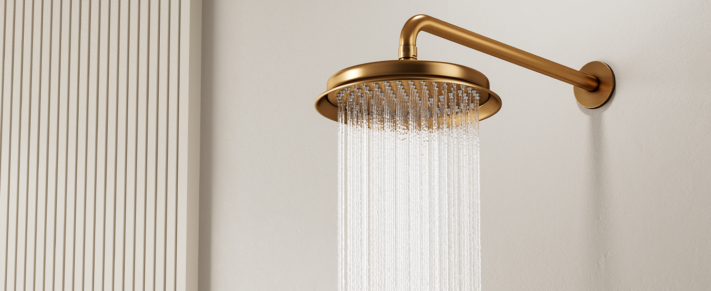 Cabezal de ducha tipo lluvia en tono dorado montado sobre un brazo de latón sobre un fondo de pared acanalado blanco. Cabezal de ducha circular con diseño de flujo de agua visible.