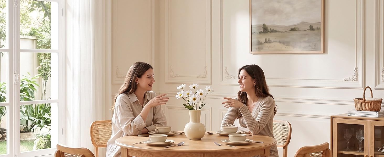 Deux femmes assises à la table à manger dans une pièce lumineuse et élégante aux murs blancs, appréciant des boissons et discutant près des fenêtres et des arrangements floraux.