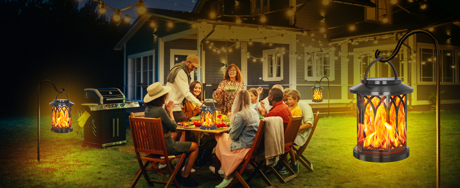 Outdoor lantern with flame-like LED lights. Metal construction with decorative cutouts. Illuminated in warm orange glow, creating ambiance for evening patio gathering in background.