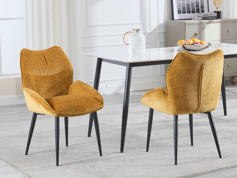 Due sedie da pranzo moderne con rivestimento in velluto giallo senape con gambe in metallo nero, esposte accanto al tavolo da pranzo bianco in un ambiente contemporaneo.