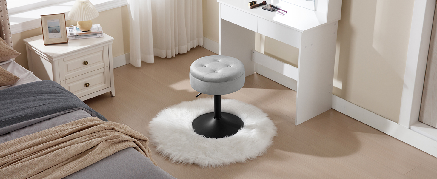 Round gray upholstered stool with a black pedestal base. Placed on a white furry rug in a bedroom setting.