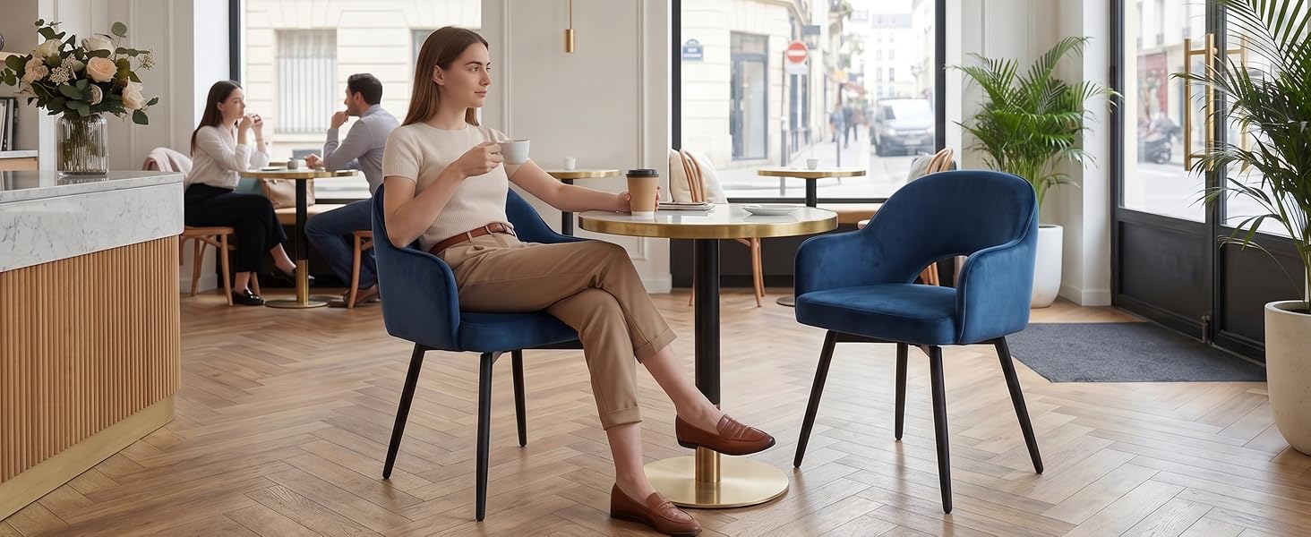 Scena da ufficio con più persone, donna in primo piano seduta su sedia blu scuro al tavolo rotondo, interni moderni con elementi in legno, skyline della città visibile attraverso le finestre.