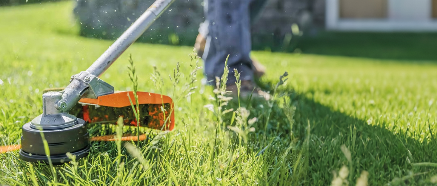 Outdoor garden trimmer with orange and black body in use on grass lawn, showing rotating cutting mechanism at ground level with person's feet visible.