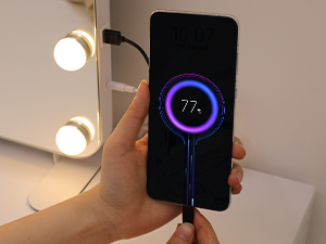 Hand holding a smartphone displaying a circular battery charging indicator showing 77% charge. A charging cable is connected to the phone.