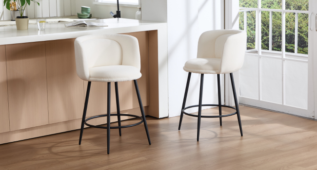 Dos taburetes de bar tapizados en color crema con patas de metal negro, que se muestran en una cocina moderna con gabinetes y ventanas de madera.