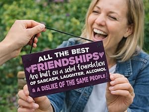 Text reads 'ALL THE BEST FRIENDSHIPS Are Built on a Foundation OF SARCASM &amp; LAUGHTER, ALCOHOL &amp; A DISLIKE OF THE SAME PEOPLE' on purple sign being held outdoors.