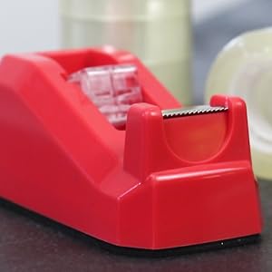 Red desktop tape dispenser with a serrated edge for cutting tape. Shown alongside clear tape rolls on a gray surface.