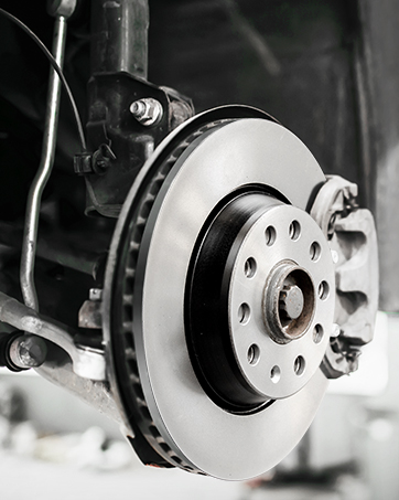 Close-up of a car brake rotor and caliper assembly. The silver disc brake rotor features ventilation holes and a smooth, reflective surface.
