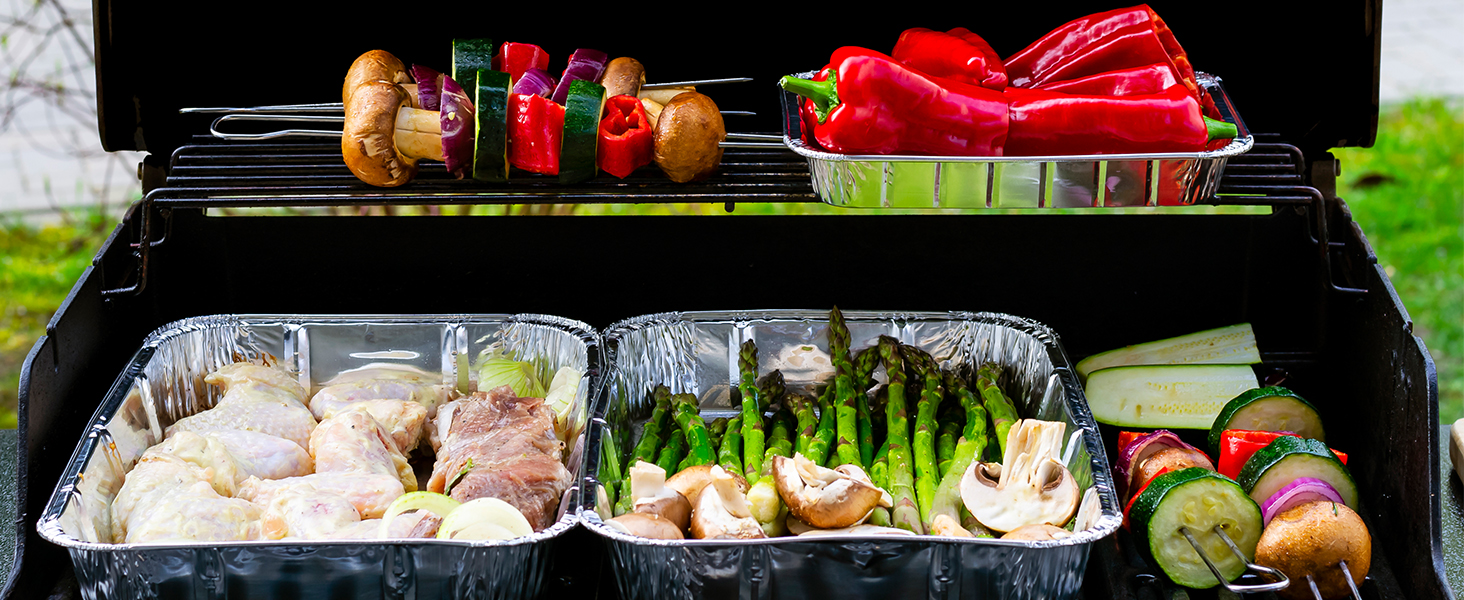 avec divers aliments. Le panier supérieur contient des brochettes de légumes. Le fond contient des plateaux en aluminium avec de la viande crue et des asperges. Poivrons rouges au premier plan.