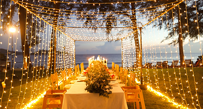 Configuration de salle à manger en plein air avec des guirlandes lumineuses, des bougies et des tables. Une pergola en bois encadre la scène avec l'océan visible en arrière-plan.