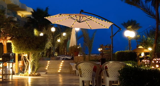 Outdoor patio umbrella with LED lights. Large white canopy illuminated at night. Set in tropical garden setting with seating visible.