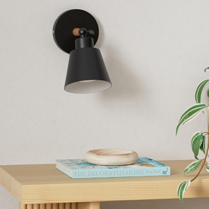 applique murale conique noire montée sur un mur blanc. Table en bois ci-dessous avec livre bleu et petite plante visible.