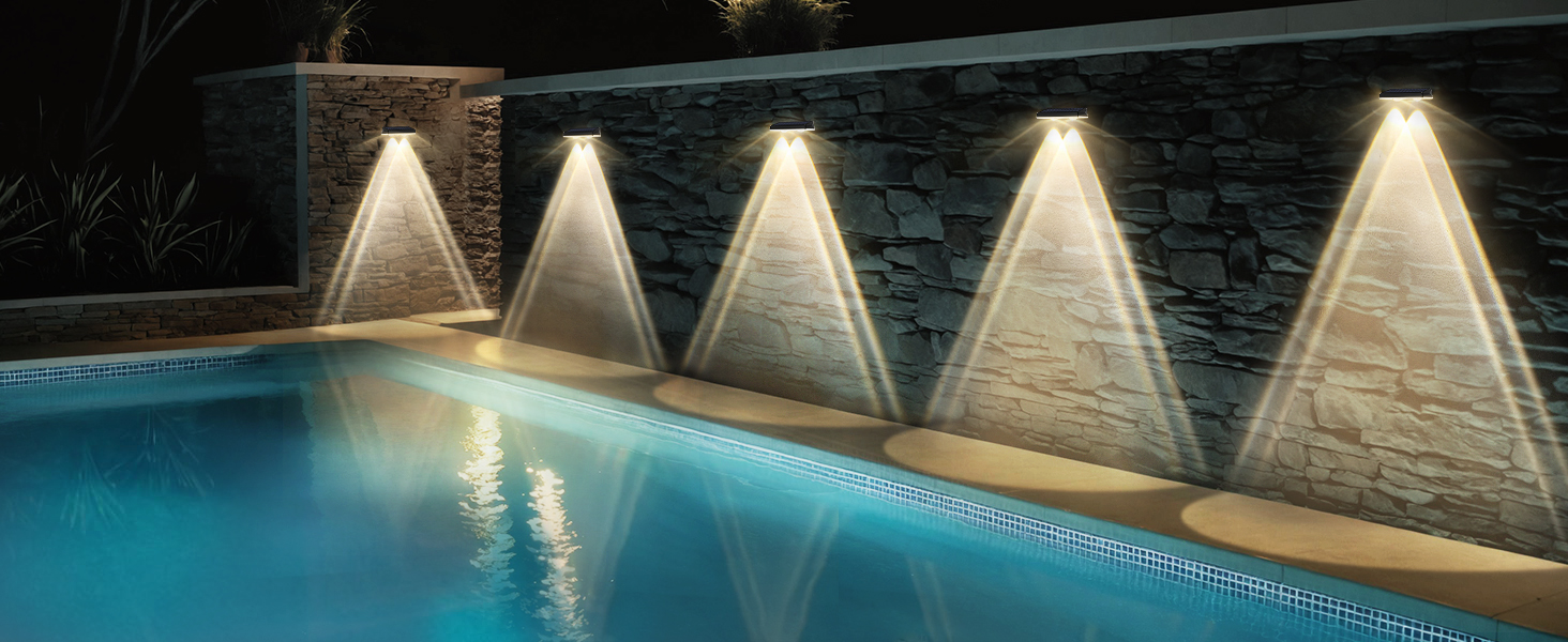Piscine extérieure illuminée la nuit. Cinq lampes murales créent des faisceaux triangulaires sur un mur de pierre, se reflétant sur la surface calme de l'eau