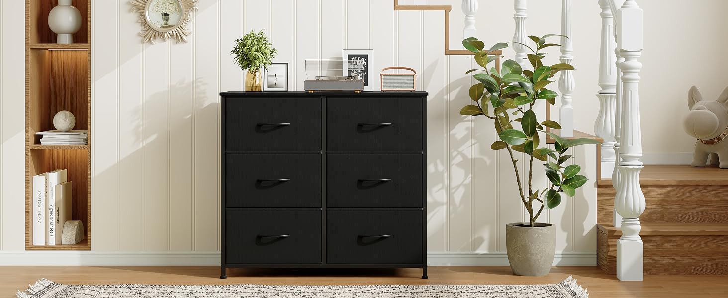 Commode en bois noire à six tiroirs, placée contre un mur lambrissé blanc. Plantes en pot et objets de décoration visibles à proximité dans un cadre domestique