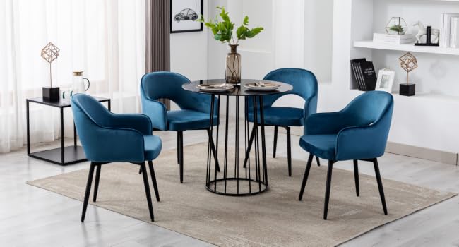 Modern dining set with round glass-top table and four blue upholstered chairs. Black metal table base. Room features white walls, decorative shelves, and potted plant.