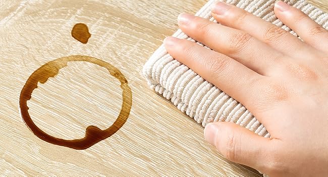 Coffee stain on light wood surface, with a hand wiping nearby using a white cloth or towel. Shows potential for cleaning or stain removal context.