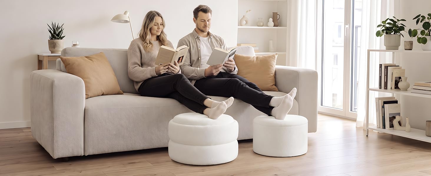 Zwei Personen sitzen auf einem hellgrauen Polstersofa mit weißen osmanischen Kissen im modernen Wohnzimmer. Helles Interieur mit Bücherregalen, Pflanzen und natürlichem Licht im Hintergrund.