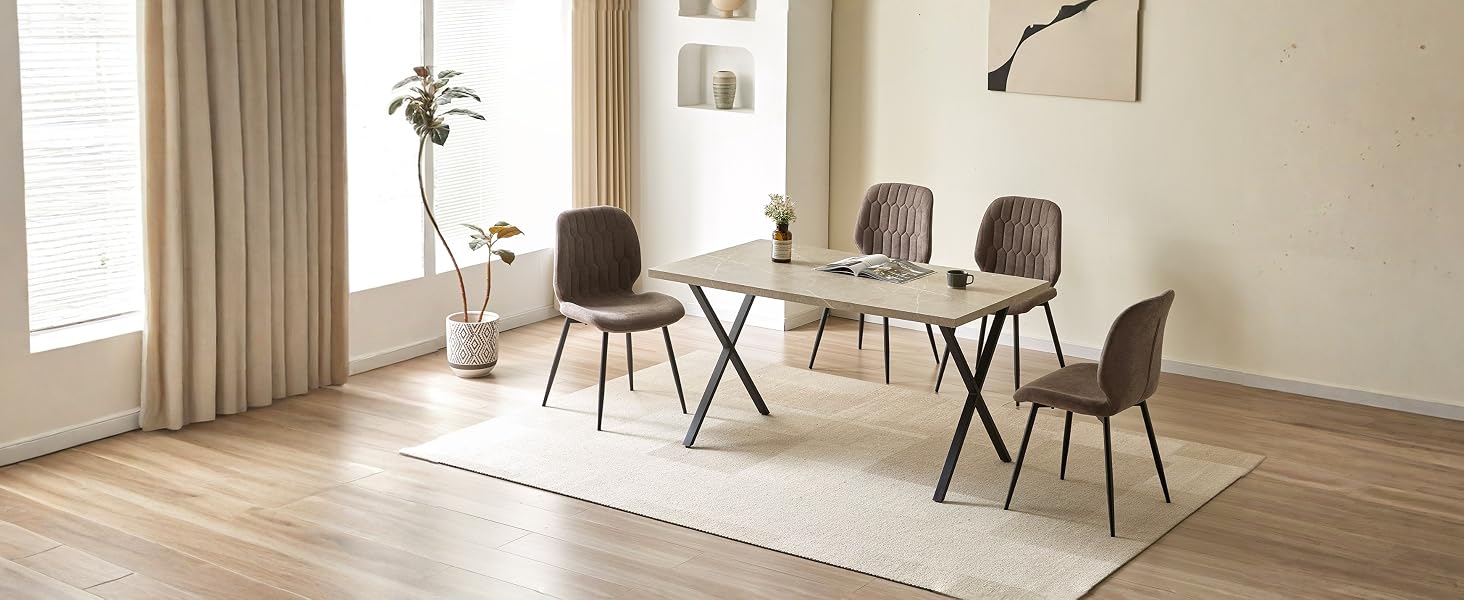 L'intérieur de la salle à manger moderne comprend une table rectangulaire en verre avec des pieds en X en métal noir, quatre chaises rembourrées brunes, un parquet en bois clair, des rideaux beiges et un tapis blanc.