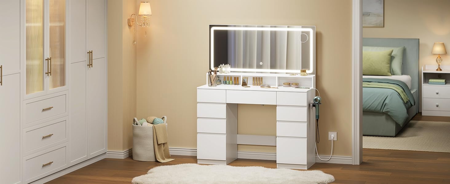 Scrivania da trucco bianca con specchio e cassetti portaoggetti posizionata nell'angolo della camera da letto con pareti beige, armadio bianco visibile a sinistra, biancheria da letto verde e opere d'arte visibili a destra.