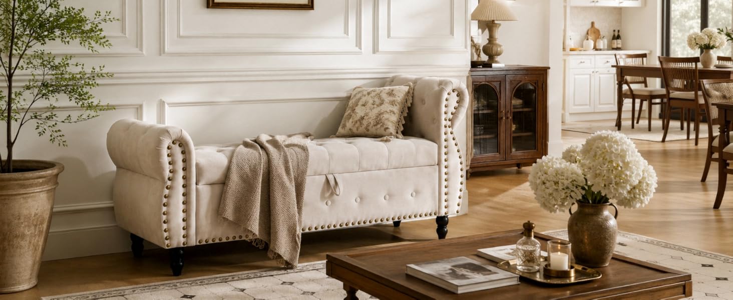 Cream-colored upholstered armchair with rolled arms and nailhead trim in furnished living room setting with side table and floral arrangement.