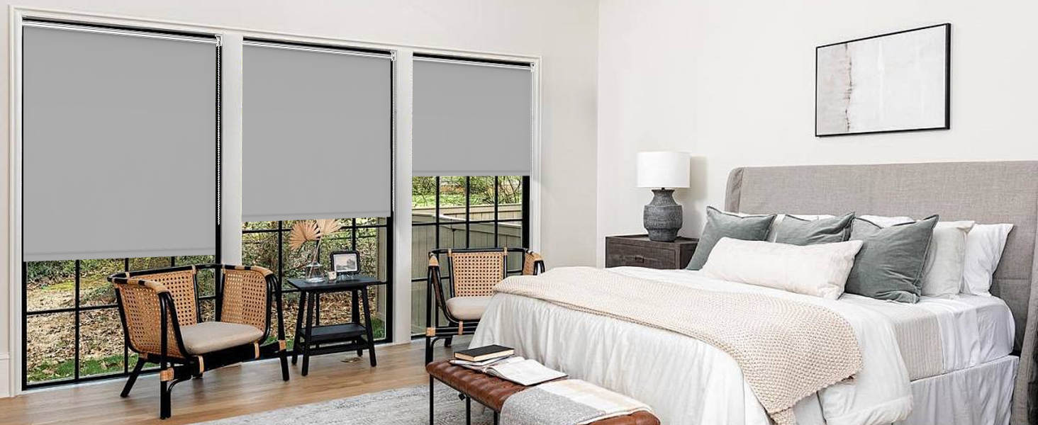 Chambre moderne avec trois grandes fenêtres dotées de stores gris. La literie blanche, la décoration neutre et les meubles en bois créent une esthétique minimaliste.