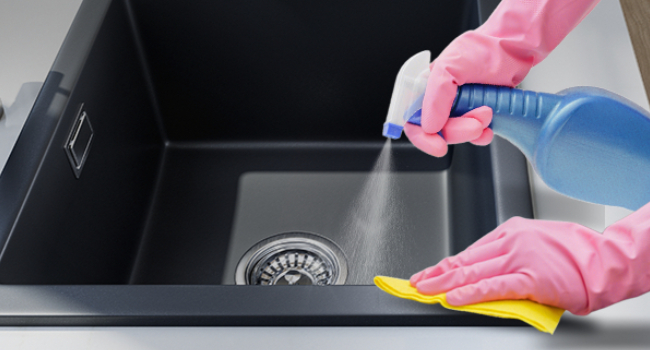 Hands in pink rubber gloves cleaning a dark-colored kitchen sink. One hand sprays cleaner from a blue bottle while the other wipes with a yellow cloth.