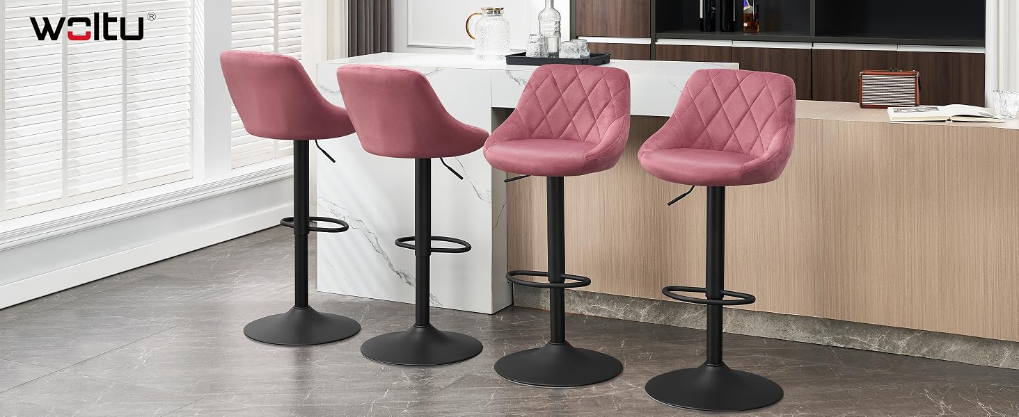 Four pink upholstered bar stools with quilted backrest design, black circular bases, and footrests. Positioned in a modern kitchen setting with wooden cabinets and white countertops.