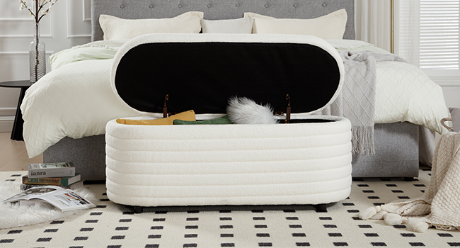 White upholstered storage ottoman with hinged lid, placed at the foot of a bed with white bedding. Ottoman interior lined with dark fabric.