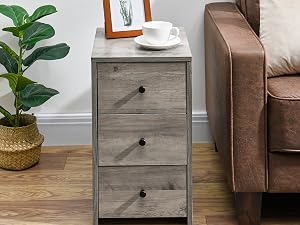 Grauer Nachttisch aus Holz mit drei Schubladen neben einer braunen Ledercouch. Dekoriert mit einer Pflanze, einem gerahmten Bild und einer Kaffeetasse
