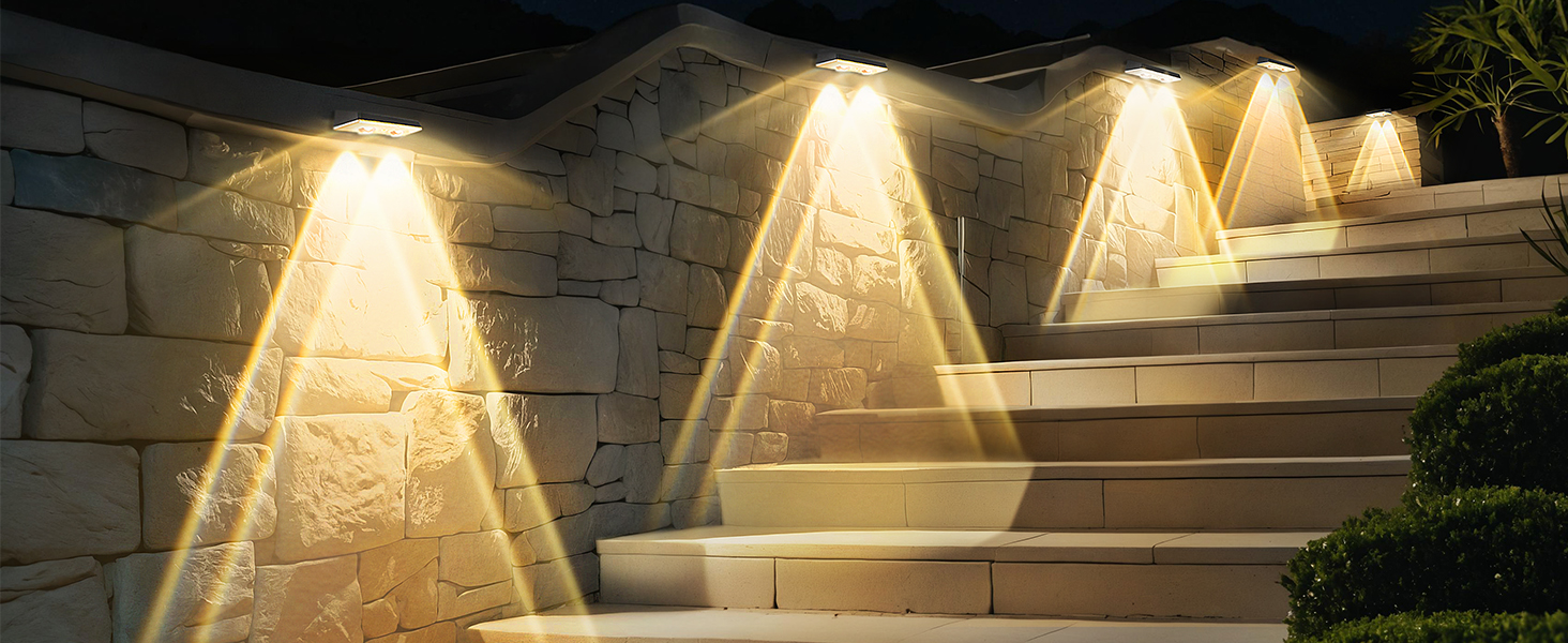 Scala esterna in pietra illuminata da applique che creano fasci triangolari di luce calda. L'illuminazione mette in risalto la texture del muro e dei gradini in pietra.