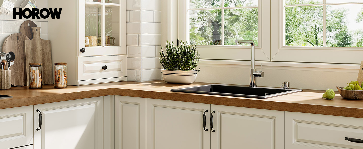 Modern white kitchen with wooden countertops, featuring a sink, windows overlooking greenery, and various kitchen accessories including a plant and cutting boards.