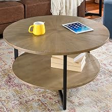 Round wooden coffee table with two tiers in a living room setting. Table displays a yellow mug, iPad, and books. Sofa, throw blanket, and patterned rug visible.
