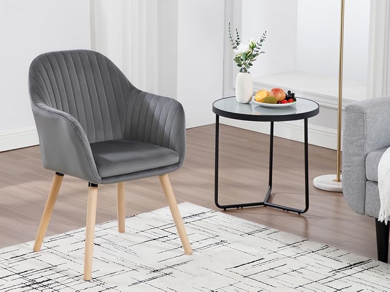 Gray upholstered armchair with wooden legs next to round side table. White rug underneath, fruit bowl on table. Modern living room setting.