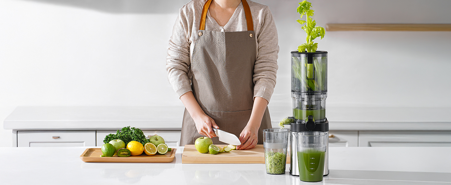 Aufrechte Saftpresse auf der Küchentheke. Person in beiger Schürze, die Obst und Gemüse zum Entsaften zubereitet. Grünes Blattgemüse, das in der Saftpresse sichtbar ist