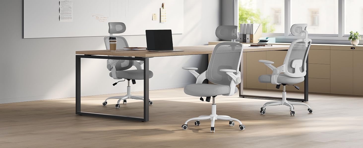 Configuración de oficina moderna con sillas grises ergonómicas, escritorio de madera y computadora portátil. Espacio lleno de luz con paredes blancas y detalles vegetales.