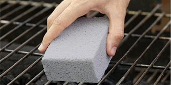 Tenez à la main un bloc de nettoyage rectangulaire gris au-dessus d'une grille. Le produit semble être conçu pour frotter et nettoyer les grilles de barbecue