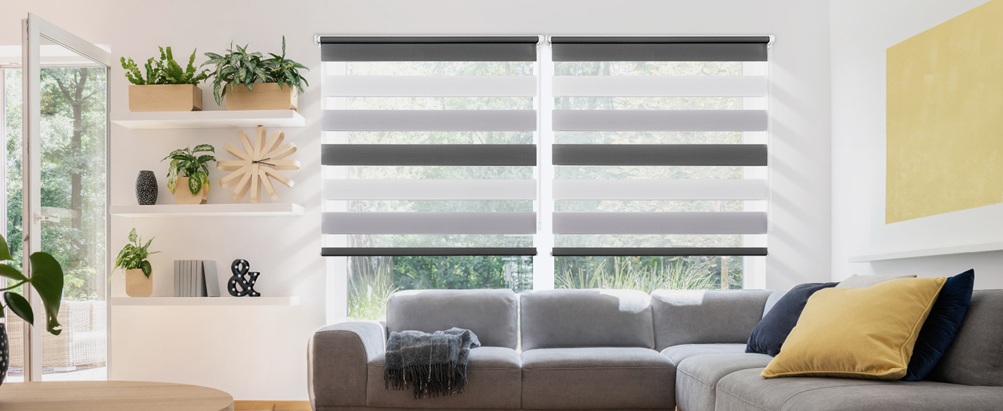 Salon moderne avec stores superposés en blanc et gris. Canapé sectionnel gris, coussin décoratif jaune et plantes sur des étagères flottantes visibles.